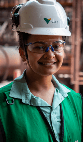 A women smiling in an operational area. She is wearing a light green shirt, a darker green vest, goggles and a white helmet with Vale logo.