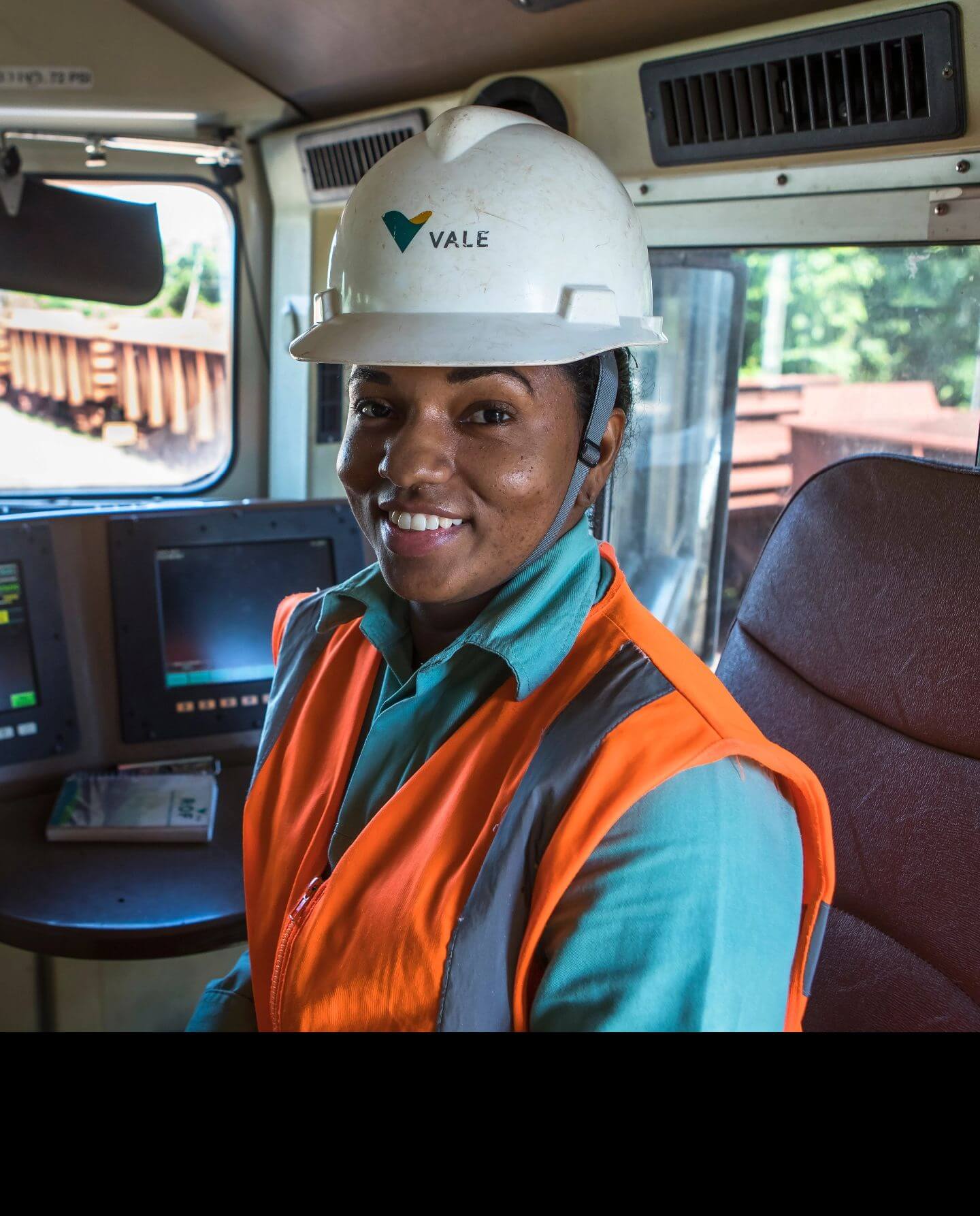 Mulher negra sorrindo dentro de um veículo. Ela usa uniforme verde da Vale, colete laranja e capacete branco com logotipo da empresa.