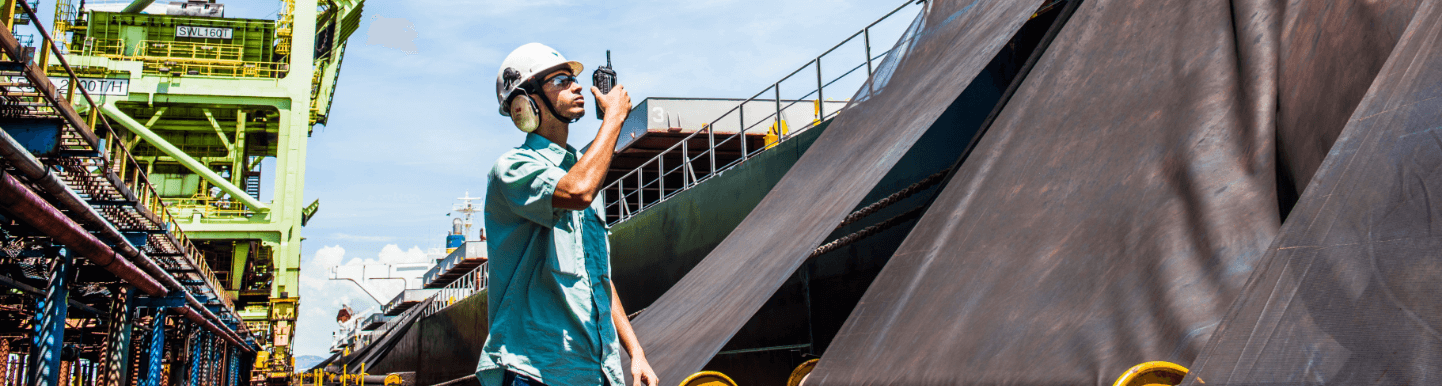 Empregado da Vale em pé no meio de uma área de operações. Ele está usando uma camisa verde clara, óculos de proteção, protetores de ouvido, capacete branco com logotipo da Vale e segura um rádio comunicador perto da boca com uma das mãos.