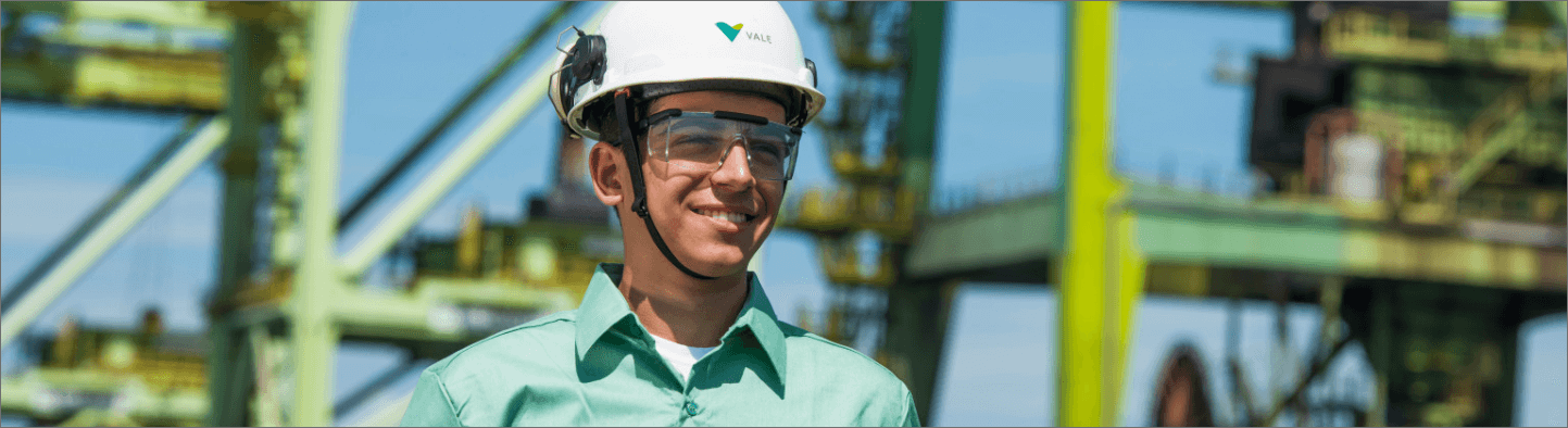 Homem sorrindo em uma área operacional. Ele usa camisa verde clara, óculos de proteção e capacete branco com logotipo da Vale.