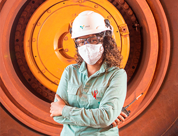 Chest to head shot of a woman. She is wearing a light green uniform, helmet, goggles, and mask. In the background is a truck wheel.