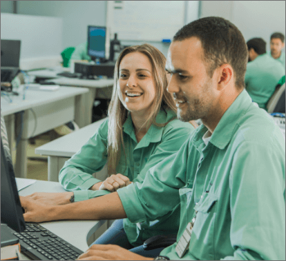 A man and a woman sitting side by side in an office. The two are wearing light green uniform shirts and looking at a computer screen. The woman is smiling. In the background, there are other desks with computers and other people.