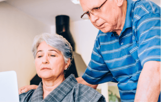 Elderly couple looking at a laptop. The woman is sitting typing and the man is standing with his hands behind her back.