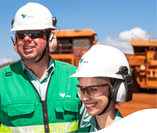 Two Vale employees – a man and a woman – smiling in an operational area with large vehicles in the background. The two are wearing green uniforms, goggles, ear muffs and white helmets with Vale logo.