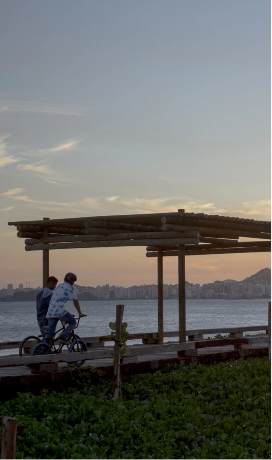Duas pessoas passeiam de bike na orla da praia. Próximo delas há uma estrutura de madeira formando uma espécie de telhado com as laterais abertas.