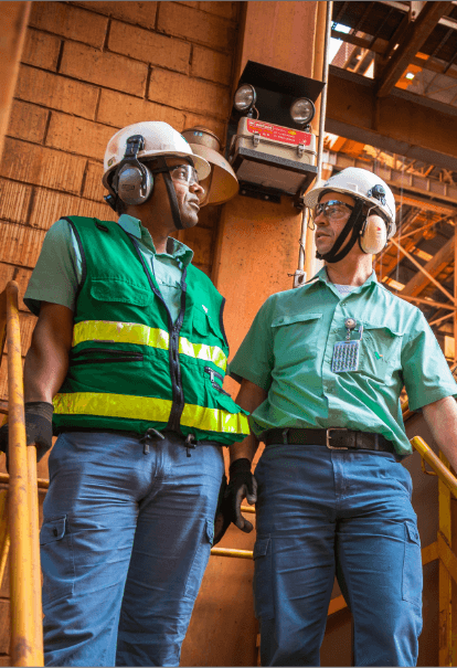 Foto de dois homens em uma área de operação conversando com estruturas de ferro ao fundo. Eles estão usando camisa de botões verde Vale, calça, capacete, óculos de proteção, luva e proteção nos ouvidos. Um deles está usando colete verde e listras fluorescentes.