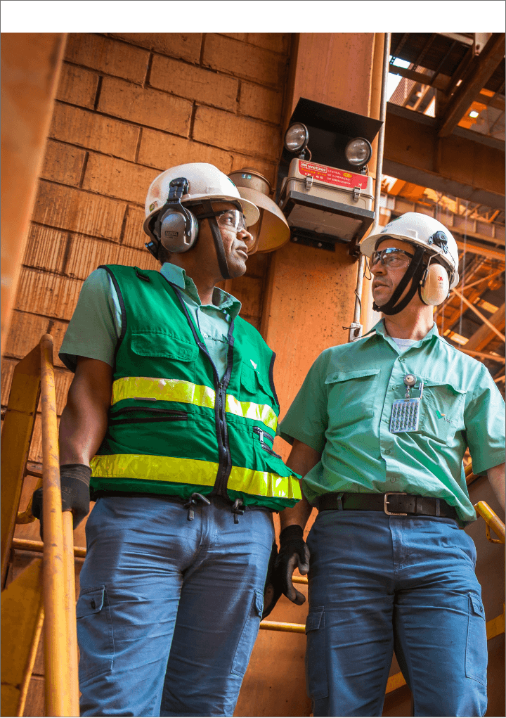 Foto de dois homens em uma área de operação conversando com estruturas de ferro ao fundo. Eles estão usando camisa de botões verde Vale, calça, capacete, óculos de proteção, luva e proteção nos ouvidos. Um deles está usando colete verde e listras fluorescentes.