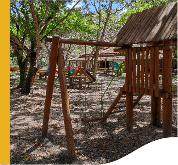 Parquinho infantil ao ar livre. Os brinquedos, como balança e escorregador, são feitos de madeira. Algumas árvores trazem sombra ao local.