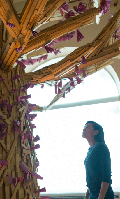 Uma mulher olha para uma escultura que se assemelha com uma árvore. A escultura é feita de pedaços de madeiras e origamis lílas.