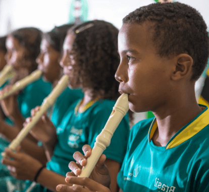 Crianças, lado a lado uma da outra, tocam flautas. Todas usam uma camiseta verde com gola amarela.