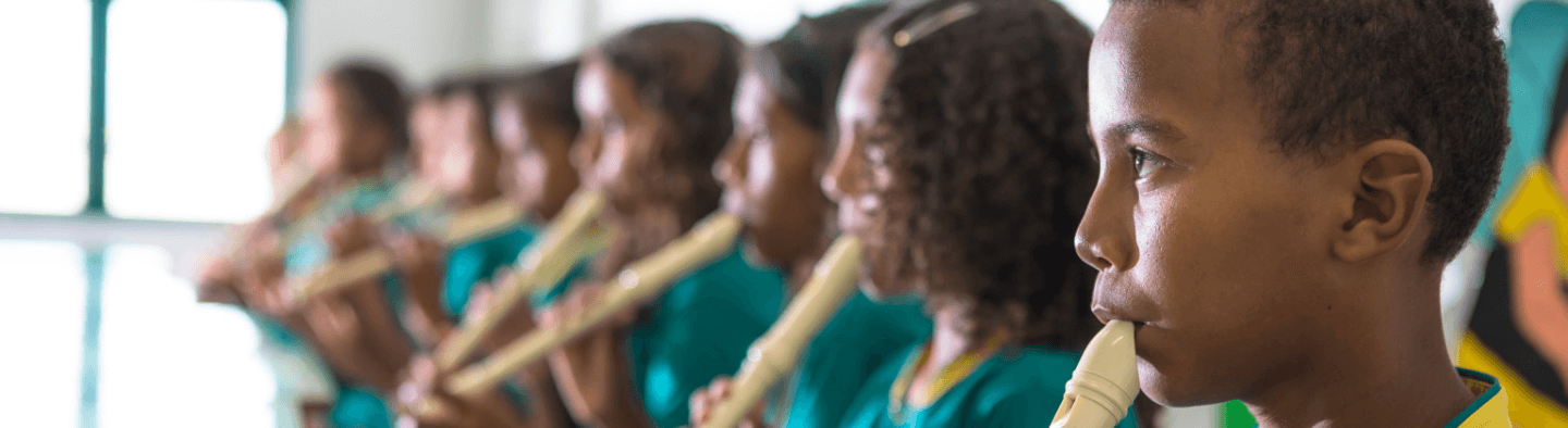 Children, side by side with each other, playing flutes. They all wear a green T-shirt with a yellow collar.