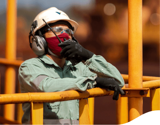 Empregado Vale, de capacete, óculos, máscara e luvas de proteção, fala em um rádio transmissor. Ele está apoiado em uma estrutura amarela de metal.