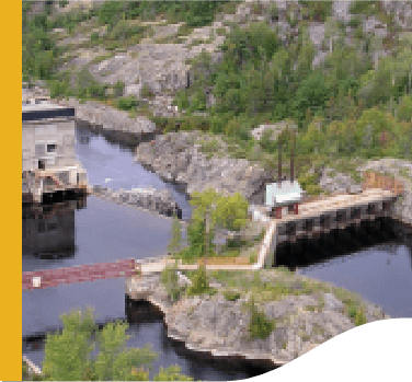 Foto da pequena central hidrelétrica Wabageshik com uma estrutura de concreto, pedras, vegetação e água em movimento passando pelas estruturas.