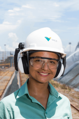 Uma empregada, ao ar livre em uma operação. Ela usa capacete e óculos de proteção, está sorrindo e de uniforme verde.