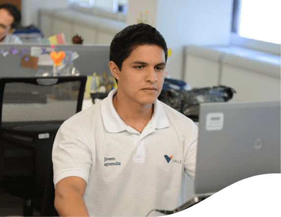 Homem sentado em um escritório olhando para um notebook que está a sua frente. Ele usa camiseta branca com o logotipo da Vale.