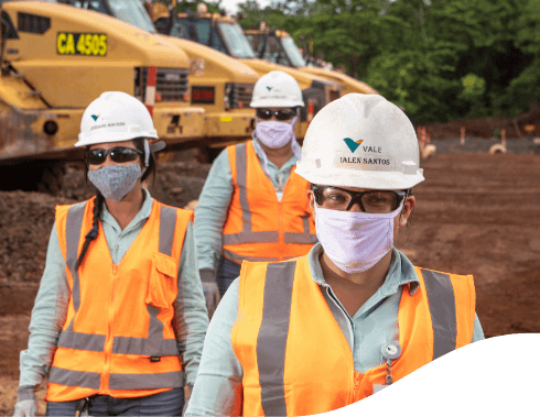 Foto da altura do peito até a cabeça de três empregados da Vale, dois home e uma mulher. Os três estão com uniforme verde da Vale, colete laranja e cinza, óculos, capacete e máscara.