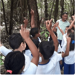 Diversas crianças, todas uniformizadas, estão em local arborizado. Ao fundo é possível ver um empregado Vale. As crianças estão com as mãos levantadas.