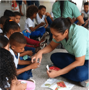 Diversas crianças estão sentadas no chão, interagindo com duas empregadas Vale.