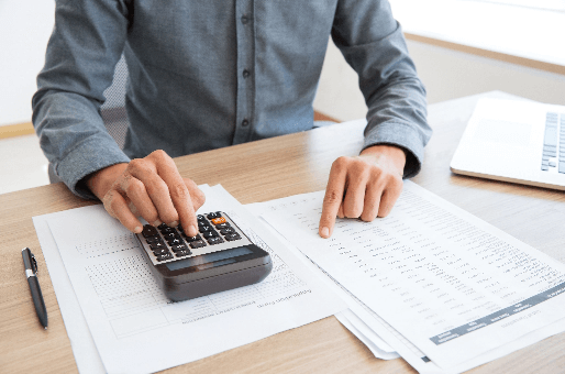 Foto tirada de cima para baixo de uma mesa com duas mãos sob ela. Uma aponta para diversos papeis e outra aperta um botão de uma calculadora.