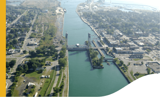 Aerial view of Port Colborne, in the corners of the image, it is possible to see a city with a lot of trees. In the middle, there is a river and a bridge connecting both sides.
