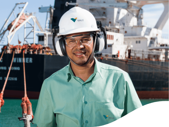 Vale employee in green uniform, helmet and goggles. He looks at the photo and smiles shyly. In the background you can see a docked ship.
