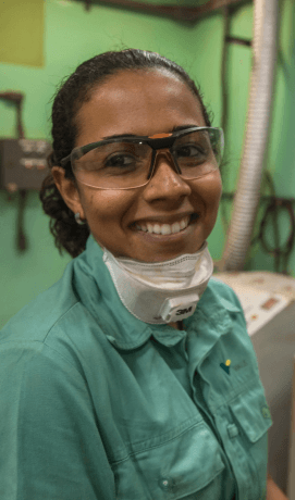 A Vale employee smiling. She is wearing a uniform, has a mask hanging around her neck, and wears goggles.