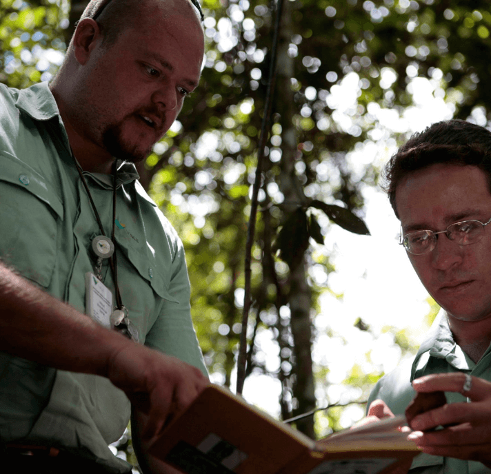 Two Vale employees are out in the open, with trees in the background. One of them is holding a notebook and both are looking toward the object.