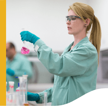 A researcher dressed in a green lab coat, goggles and gloves analyzes a test tube with pink liquid.