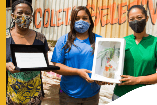 Três mulheres, todas de máscaras de proteção, pousam para foto. Uma delas segura um certificado e as outras duas seguram uma caixa com uma planta dentro.