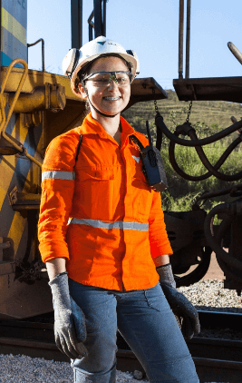 A woman is in an open space and a truck is behind her. She is wearing jeans, an orange shirt, goggles and a white helmet