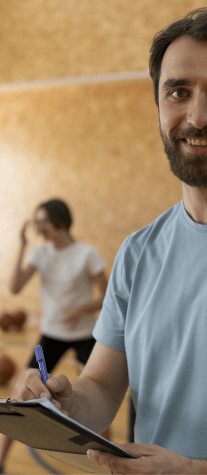 Man smiling at camera, holding a clipboard and a pen. He has a beard and short, straight, brown hair. Behind him, there are two people doing physical exercises.