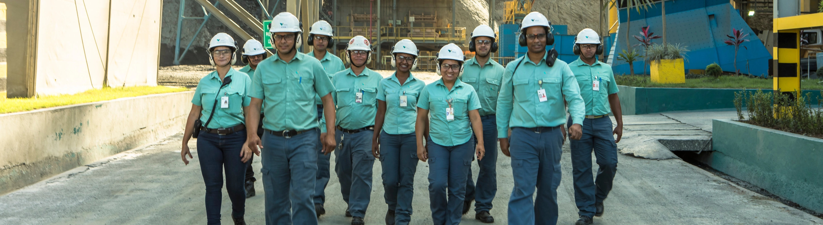 Full body photo of 10 Vale employees with an operation in the background, wearing uniforms: pants, green button-up shirt, helmet, goggles, ear protection and badge
