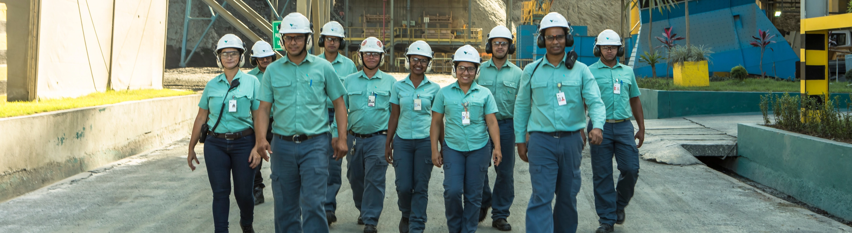 Foto de corpo todo de 10 empregados da Vale com uma operação ao fundo, usando uniforme: calça, camisa botões verde, capacete, óculos, proteção nos ouvidos e crachá