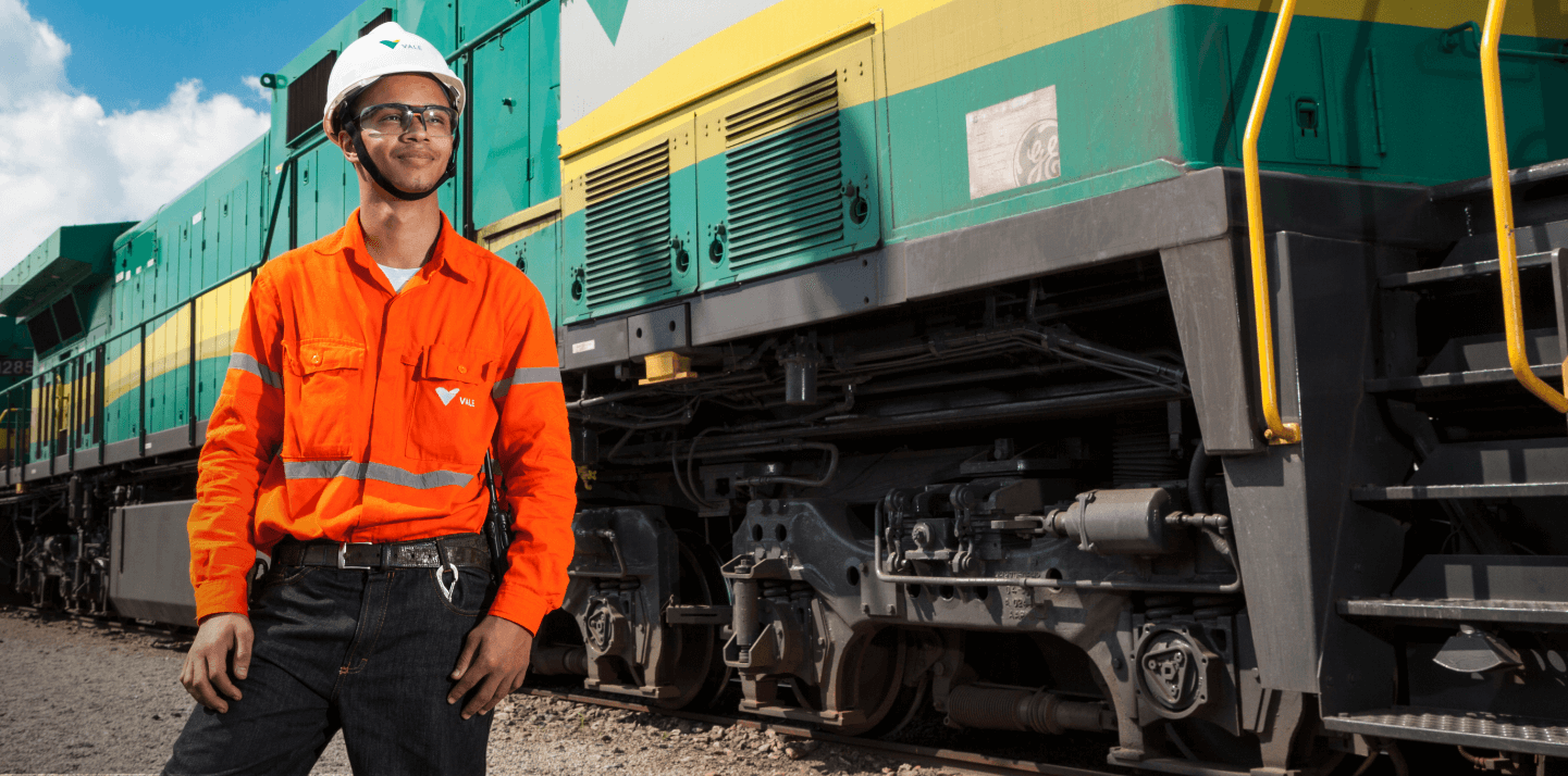 Homem parado ao lado de um trem verde, amarelo e cinza. Ele usa uniforme laranja com logotipo da Vale, óculos de proteção e capacete branco também com logotipo da empresa.