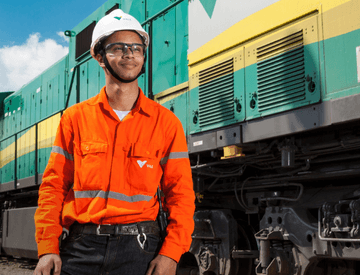 Homem parado ao lado de um trem verde, amarelo e cinza. Ele usa uniforme laranja com logotipo da Vale, óculos de proteção e capacete branco também com logotipo da empresa.