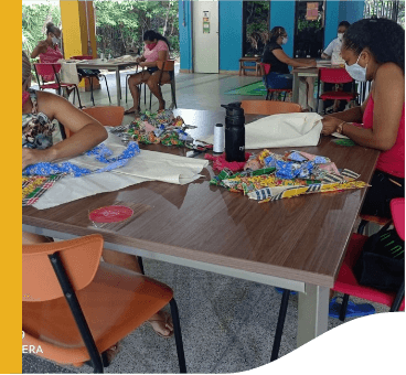 Diversas mulheres estão sentadas em volta de mesas, manuseando tecidos.