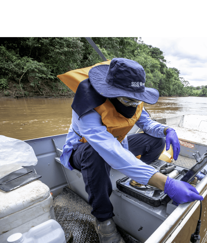 Um homem, de chapéu, máscara, óculos protetor, luvas e colete, está dentro de um barco com diversos galões e uma maleta com equipamentos.