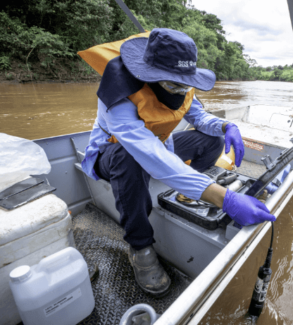 A man, wearing a hat, a mask, goggles, gloves and a vest, is inside a boat with several gallons and a case with equipment.