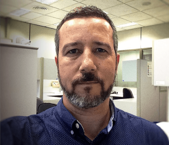 A man with short hair and a beard is in an office. His face is serious and is wearing a dark blue shirt.