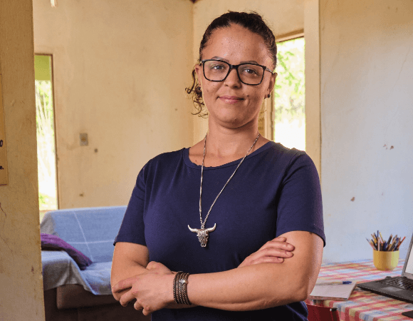 Portrait of Roziane Duarte smiling at the camera. She has her arms crossed, she is wearing glasses, a big necklace and her hair is tied up.