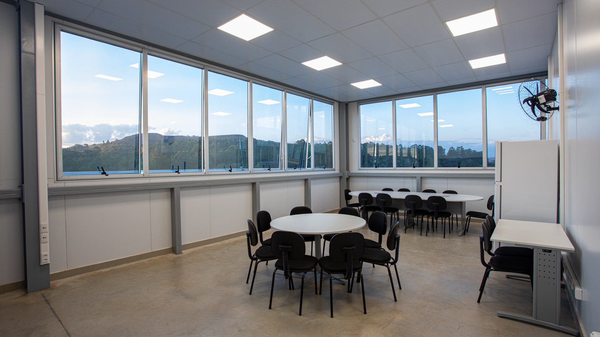 Sala com janelas ampplas e mesas redondas com diversas cadeiras ao redor. Há também um geladeira e um ventilador.