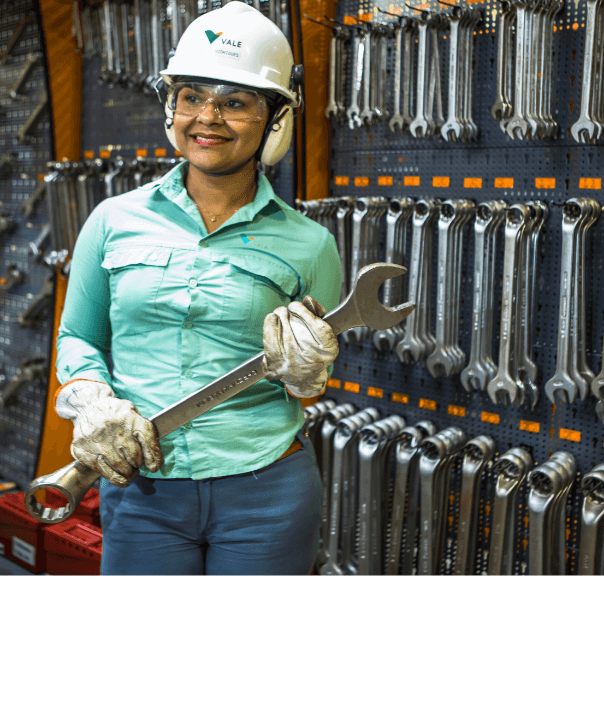 Mulher parada, segurando uma ferramenta de aço em um espaço repleto de outras ferramentas. Ela usa camisa verde clara de uniforme, luvas, óculos de proteção, protetores de ouvido e um capacete branco com logotipo da Vale.