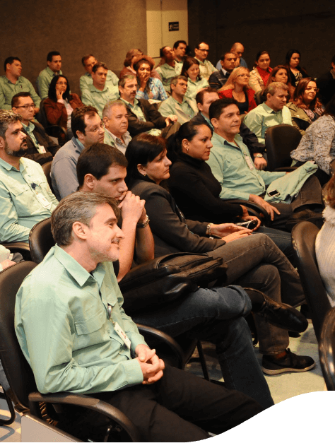 Foto de várias pessoas sentadas em uma sala em um seminário. As pessoas estão utilizando camisa verde vale de botões e calça jeans.