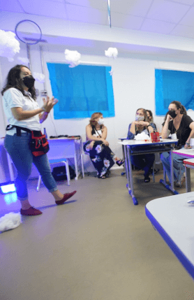 Uma mulher está de pé em uma sala de aula, enquanto outras mulheres estão sentadas olhando para ela. Todas estão de máscara.
