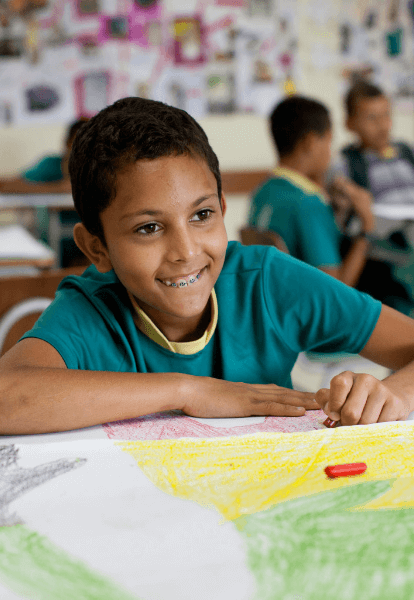 Um menino, dentro de uma sala de aula, pinta uma folha com giz de cera. Ele usa aparelho nos dentes e veste um uniforme verde e amarelo.