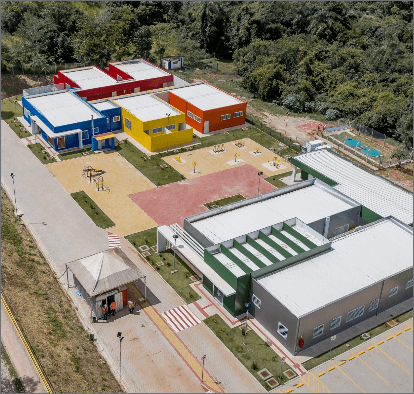 Imagem aérea de um local que se assemelha a uma escola. Há estruturas coloridas de concreto, um espaço de um parquinho e, em volta, muita vegetação.