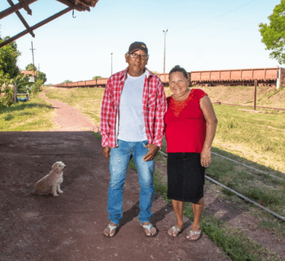 Image of an old man and an old woman. The two are standing, smiling for the photo. In the background, it is possible to see a grass area and a train.