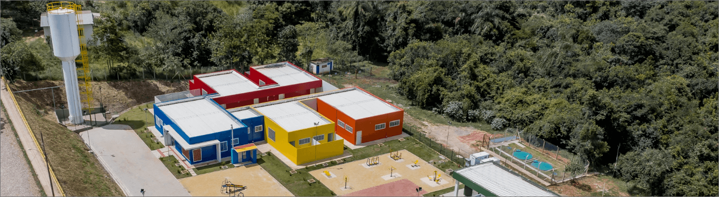 Imagem aérea de um local que se assemelha a uma escola. Há estruturas coloridas de concreto, um espaço de um parquinho e, em volta, muita vegetação.