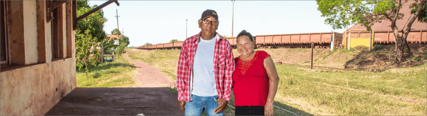 Image of an old man and an old woman. The two are standing, smiling for the photo. In the background, it is possible to see a grass area and a train.