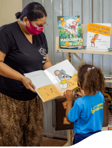 Uma mulher, de máscara e óculos, segura um livro em direção a uma menina que presta atenção na figura desenhada na página. Ao fundo há uma estante com outros livros.